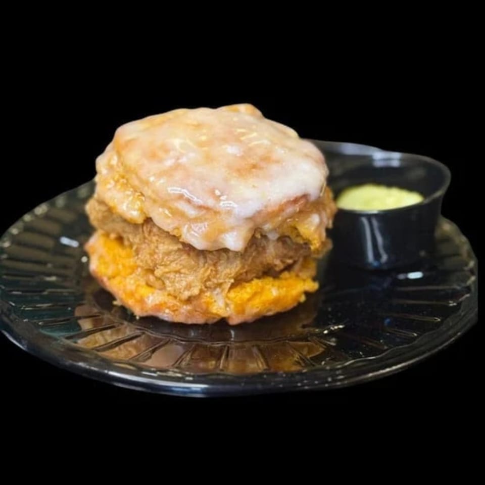 A glazed biscuit sandwich with fried chicken and a side of green sauce on a black plate.