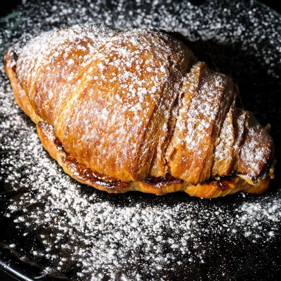 A golden-brown croissant, generously dusted with powdered sugar, sits on a dark surface.